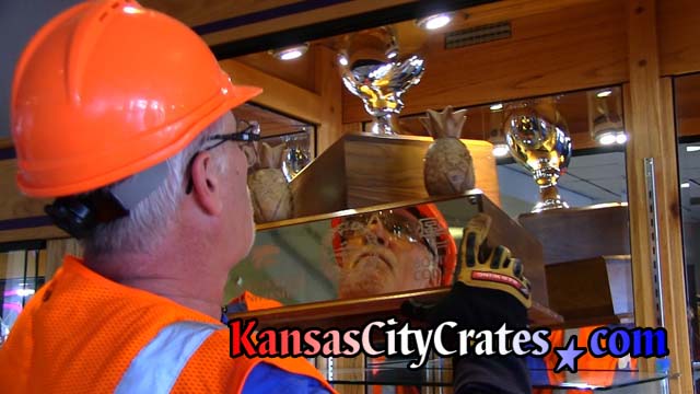 Glass cabinet holding Governor's Cup trophy opened bu crate packers wearing hard hats, gloves, eye protection, safety vests and steel toe boots